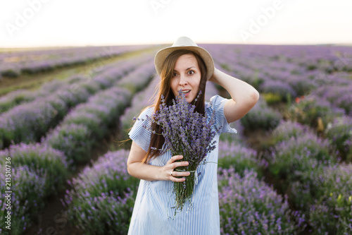 Young sensual beautiful woman in blue dress, hat on purple lavender flower blossom meadow field outdoors on summer nature background. Tender female near flowering bush with bouquet. Lifestyle concept.