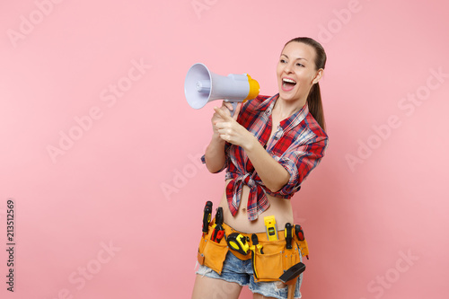 Young excited handyman woman in plaid shirt, denim shorts, kit tools belt full of variety instruments hold megaphone, screaming isolated on pink background. Female doing male work. Renovation concept.