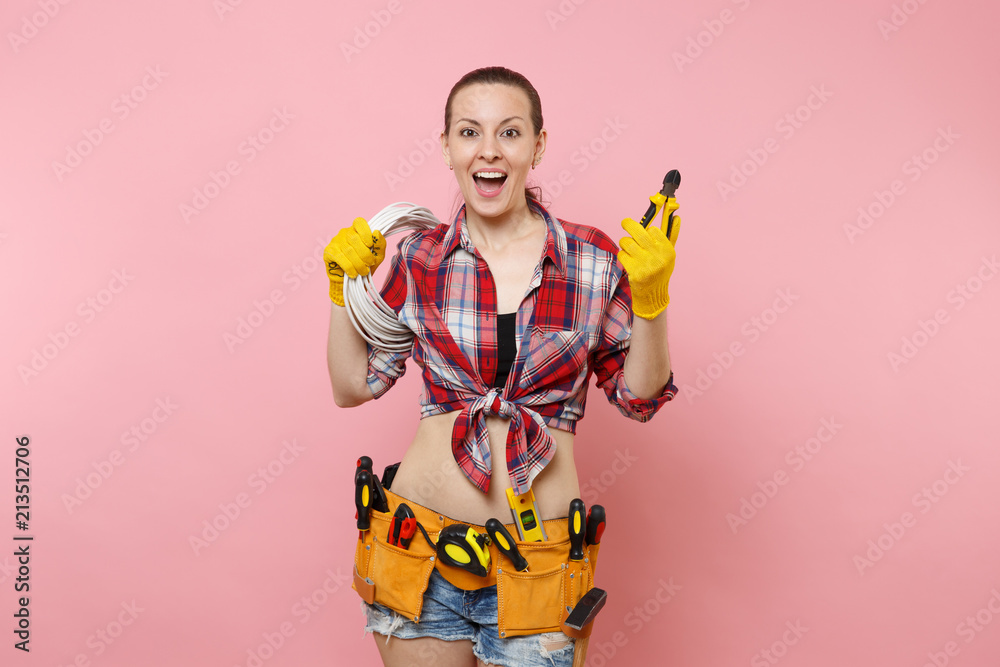 Young excited handyman woman in plaid shirt, denim shorts, kit tools belt full of variety instruments holding electric power cabel isolated on pink background. Female in male work. Renovation concept. Young excited handyman woman in plaid shirt, denim shorts, kit tools belt full of variety instruments holding electric power cabel isolated on pink background. Female in male work. Renovation concept.