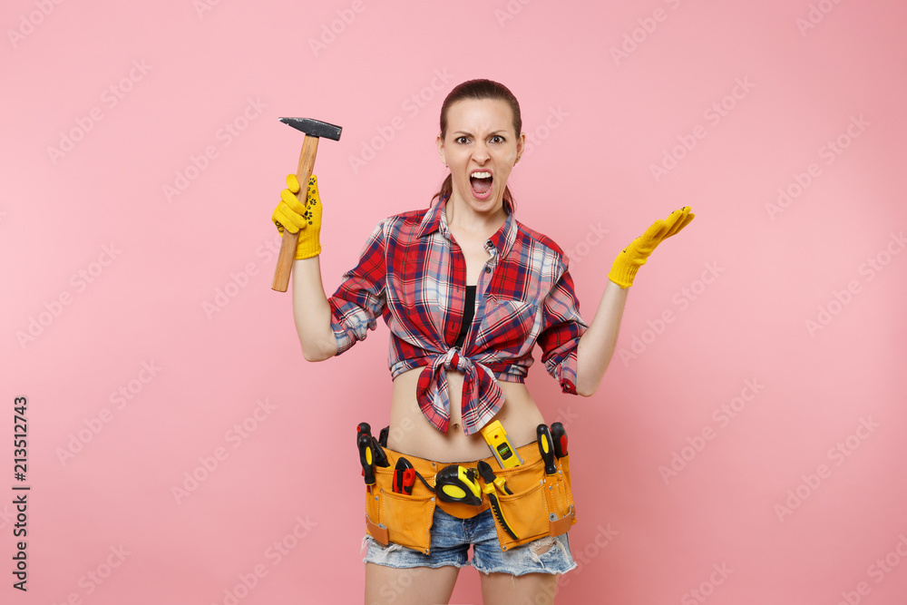 Strong young handyman woman wearing plaid shirt, denim shorts, kit tools belt full of different instruments hammer isolated on pink background. Female in male work. Renovation and occupation concept.