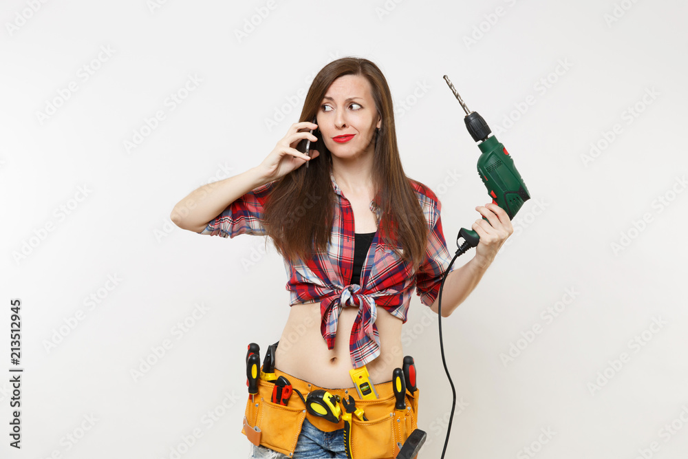 Young energy handyman woman in plaid shirt, kit tools belt full of instruments talking on mobile phone, hold power electric drill isolated on white background. Female in male work. Renovation concept. Young energy handyman woman in plaid shirt, kit tools belt full of instruments talking on mobile phone, hold power electric drill isolated on white background. Female in male work. Renovation concept.