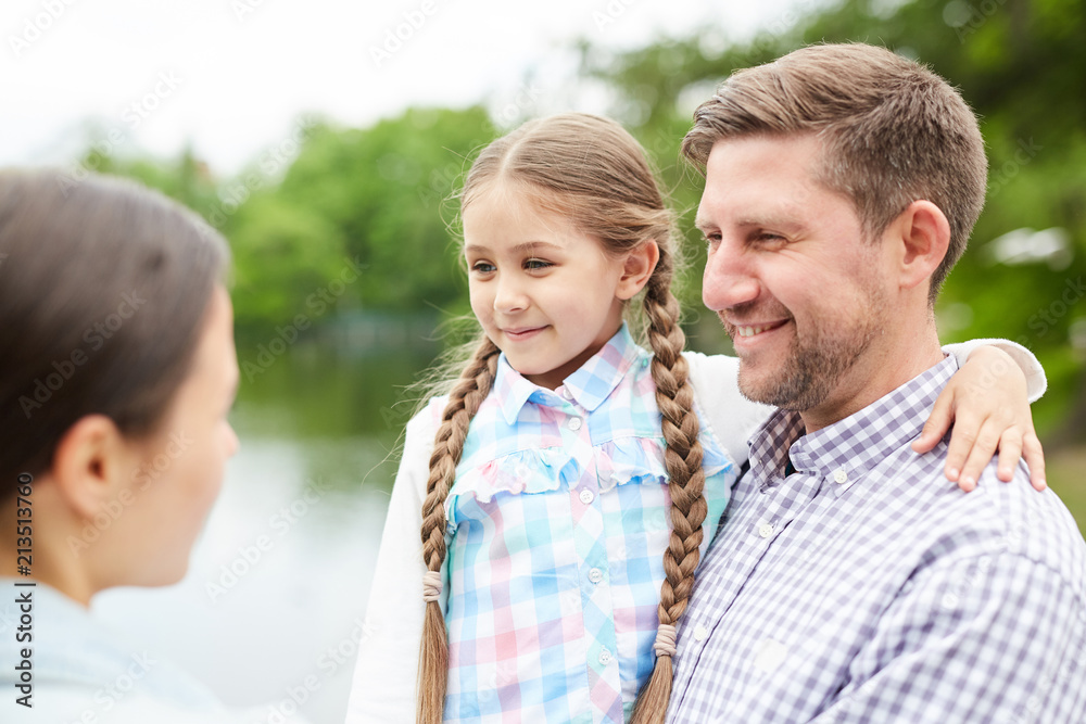 Happy young man with daughter on hands talking to woman during relax in park by waterside Happy young man with daughter on hands talking to woman during relax in park by waterside