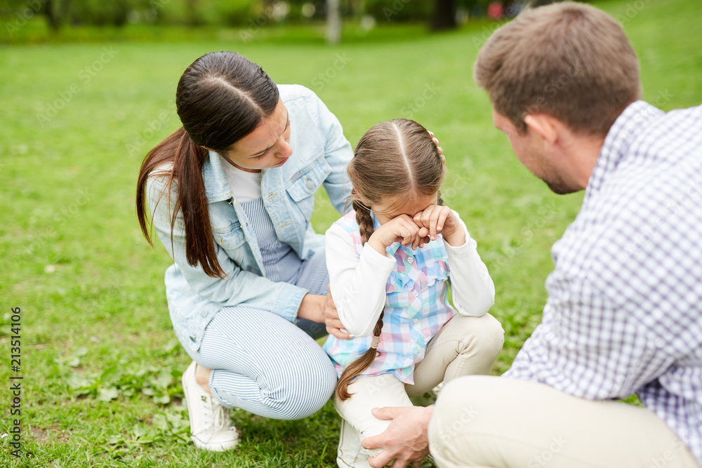 Little girl crying while sitting on grass and her parents comforting her during weekend chill
