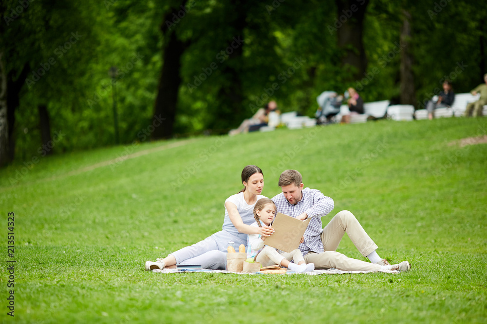 Little girl and her parents looking into open book and discussing story while relaxing at picnic on green lawn Little girl and her parents looking into open book and discussing story while relaxing at picnic on green lawn