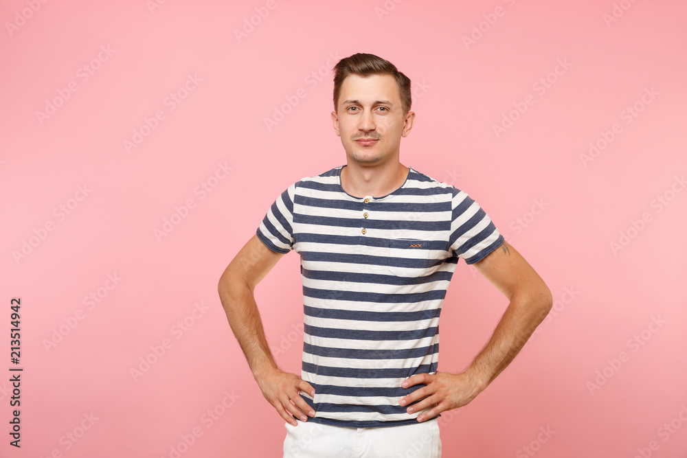 Portrait of smiling young hipster man in striped t-shirt standing with hands akimbo isolated on trending pink background. People sincere emotions, youth lifestyle concept. Advertising area copy space. Portrait of smiling young hipster man in striped t-shirt standing with hands akimbo isolated on trending pink background. People sincere emotions, youth lifestyle concept. Advertising area copy space.