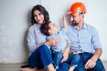happy family handsome man in helmet hardhat and charming woman and their cute little son sitting on the floor against a white wall . concept of buying repairs house ( apartment ) by young family