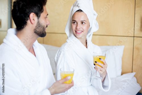 Young couple with glasses of orange or peach juice having talk in bedroom or hotel room on vacation