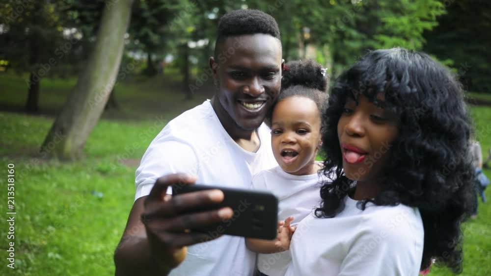 Good-looking young man holding smartphone in hand. Happy African family looking at camera of smartphone, smiling and grimacing. Daytime. Weekend. Outdoors. Summertime. Good-looking young man holding smartphone in hand. Happy African family looking at camera of smartphone, smiling and grimacing. Daytime. Weekend. Outdoors. Summertime.