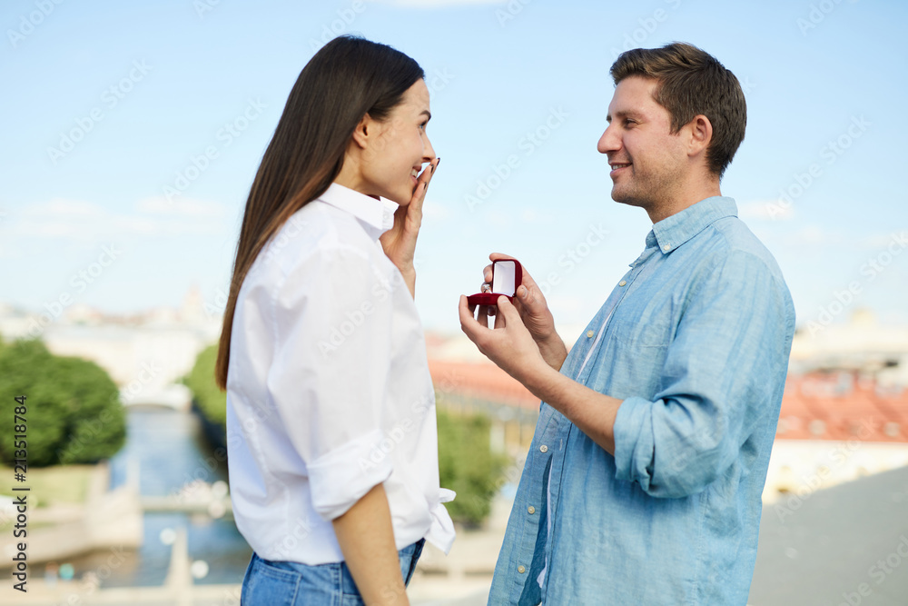Surprised girl looking at her boyfriend with open velvet box making her proposal in romantic atmosphere Surprised girl looking at her boyfriend with open velvet box making her proposal in romantic atmosphere