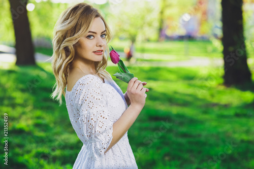 Young beautiful blonde girl in white dress with a flower tulip on a background of green grass