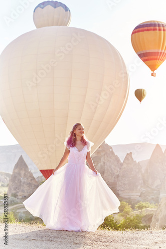 Woman in a long dress on background of balloons in Cappadocia. Girl with flowers hands stands on a hill and looks at a large number of flying balloons. Turkey Cappadocia fairytale scenery of mountains
