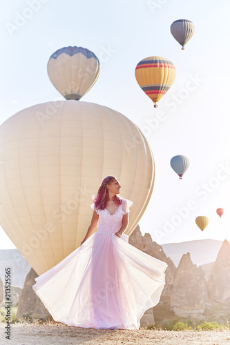 Woman in a long dress on background of balloons in Cappadocia. Girl with flowers hands stands on a hill and looks at a large number of flying balloons. Turkey Cappadocia fairytale scenery of mountains