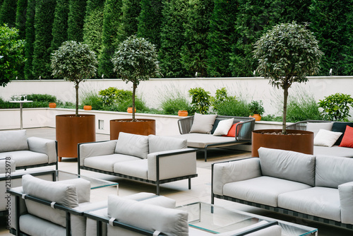 Beautiful, empty terrace with white sofas and glass tables, surrounded with green trees and flowerpots.