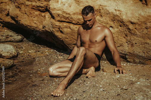 Strong face sport sexy male model wearing wet cloathes on the sea shore background with sand and water on the beach on the sunset photo