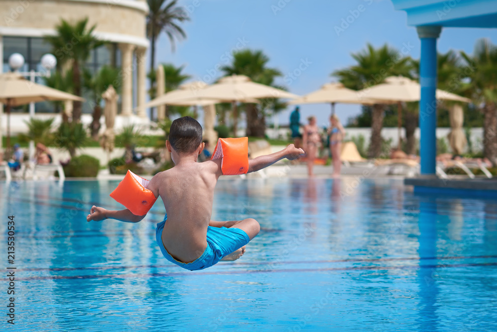 Caucasian boy in floating sleeves jumping into water in swimming pool at resort. Back view.