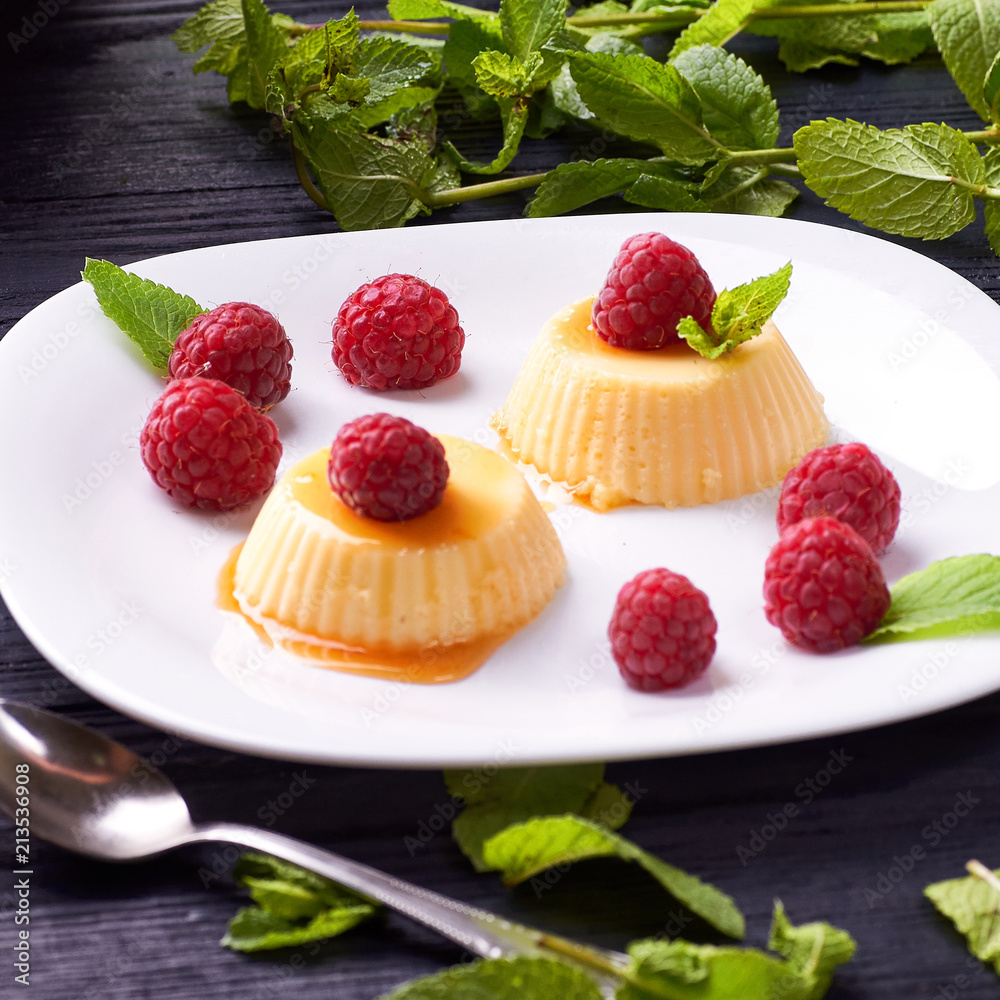 Small portioned panna cotta with fresh raspberries. Milk creamy dessert, decorated with leaves of fragrant fresh mint. White dessert plate on a black wooden table. Small portioned panna cotta with fresh raspberries. Milk creamy dessert, decorated with leaves of fragrant fresh mint. White dessert plate on a black wooden table.