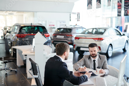 Bearded young male customer signing a contract for rent a car while sitting at car showroom
