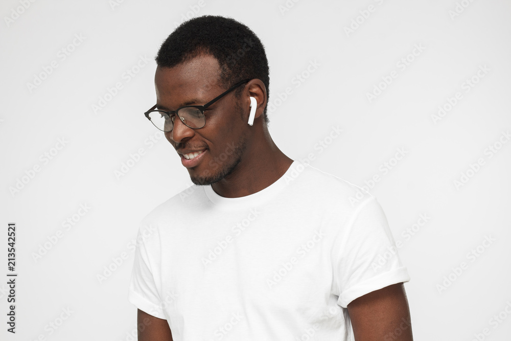 Closeup picture of handsome African man standing isolated on gray background looking down through eyeglasses having trendy earbuds without cord on enjoying music and thinking some of his thoughts Closeup picture of handsome African man standing isolated on gray background looking down through eyeglasses having trendy earbuds without cord on enjoying music and thinking some of his thoughts