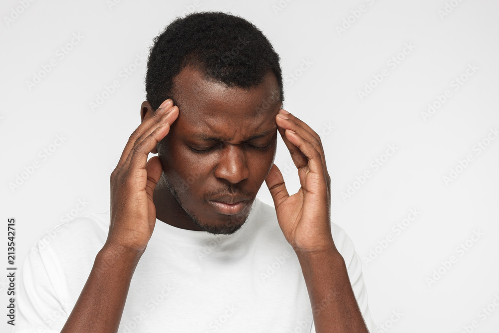 Indoor closeup of African American man pictured on gray background dressed in casual T-shirt suffering from terrible pain in head, showing it in facial expression looking helpless and miserable Indoor closeup of African American man pictured on gray background dressed in casual T-shirt suffering from terrible pain in head, showing it in facial expression looking helpless and miserable