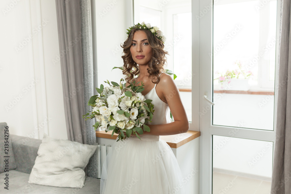 The bride in a beautiful wedding dress with a long curly hair stands in the bedroom and looks at the camera. Bride indoors with white flowers bouquet and tender wreath hairstyle
