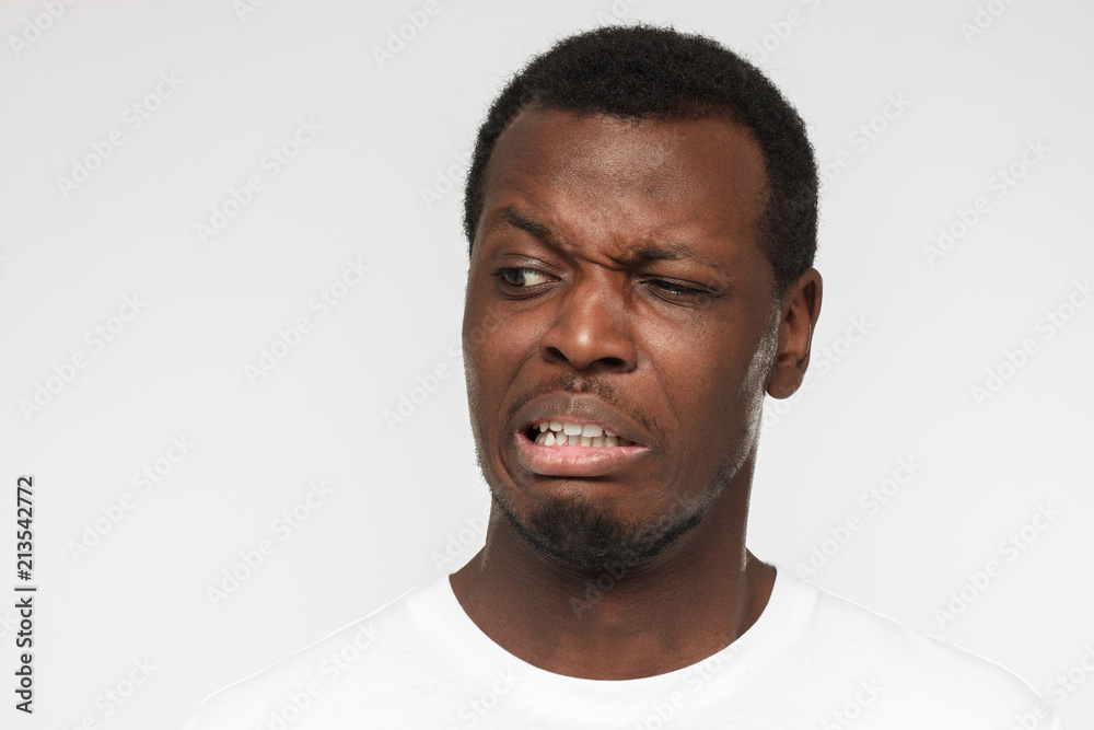 Studio photo of African American man dressed in casual T-shirt standing against grey background frowning and grinning with expression of strong disgust, absolutely dissatisfied with what he perceives Studio photo of African American man dressed in casual T-shirt standing against grey background frowning and grinning with expression of strong disgust, absolutely dissatisfied with what he perceives