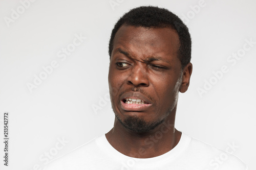 Studio photo of African American man dressed in casual T-shirt standing against grey background frowning and grinning with expression of strong disgust, absolutely dissatisfied with what he perceives