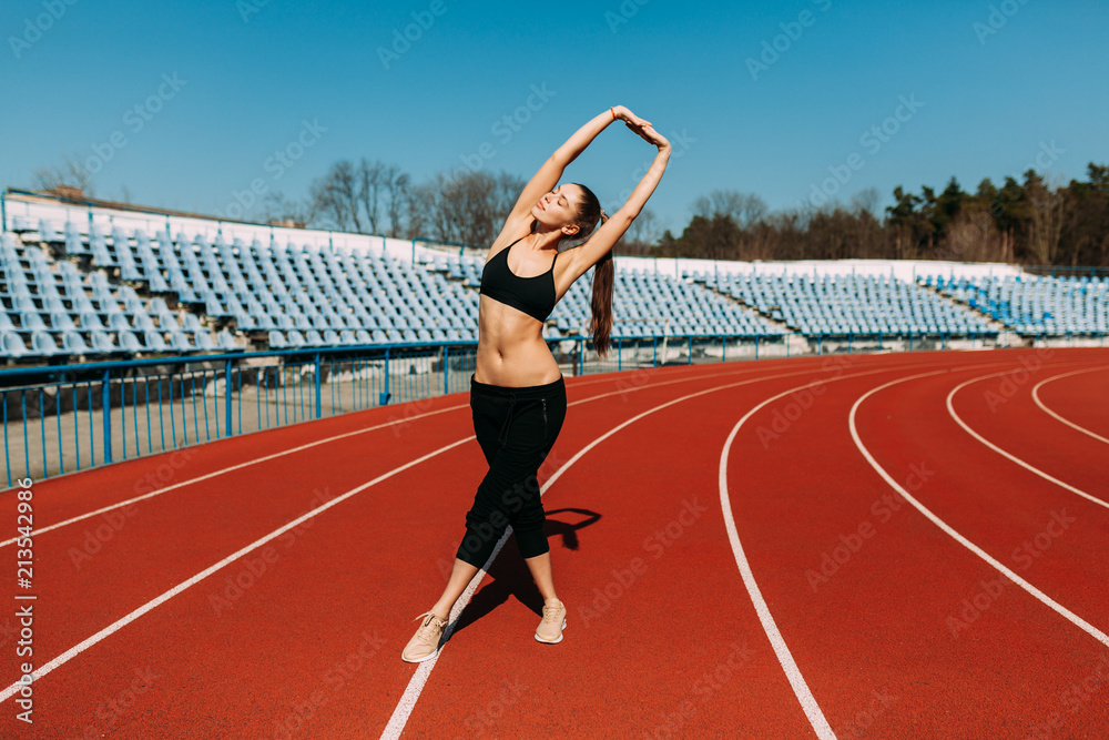 Fototapeta premium A beautiful sports girl in black sportswear is doing a warm-up before practicing sports at the stadium. The concept of a healthy lifestyle