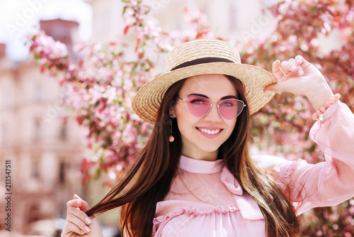 Outdoor close up portrait of young beautiful happy smiling woman wearing stylish pink cat eye sunglasses, straw hat, earrings, blouse, posing in street of city. Copy, empty space for text