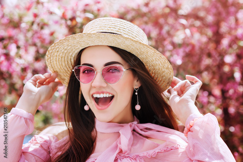 Outdoor close up portrait of young beautiful happy smiling woman wearing straw hat, pink sunglasses, earrings, blouse, posing street, near blooming tree. Advertisement concept