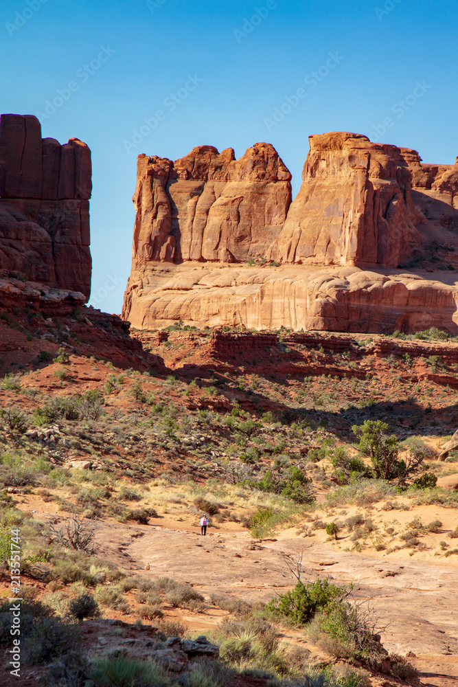 Fototapeta premium Looking back up the wash at the monumental rock formation that forms the Park Avenue Trail in Arches National Park totally dwarfs the lone hiker