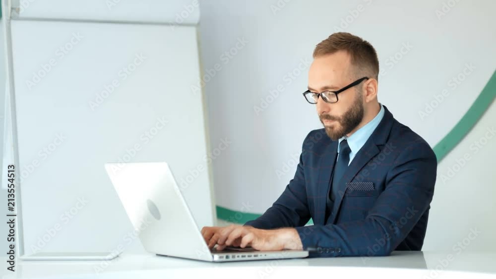 Attractive businessman in suit and glasses working with laptop in white office Attractive businessman in suit and glasses working with laptop in white office