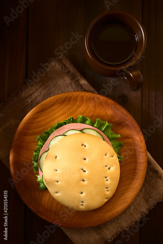 Traditional Chilean Hallulla bread roll prepared as a sandwich with lettuce, cold cut and cucumber served on wooden plate with a cup of tea, photographed overhead on dark wood with natural light photo