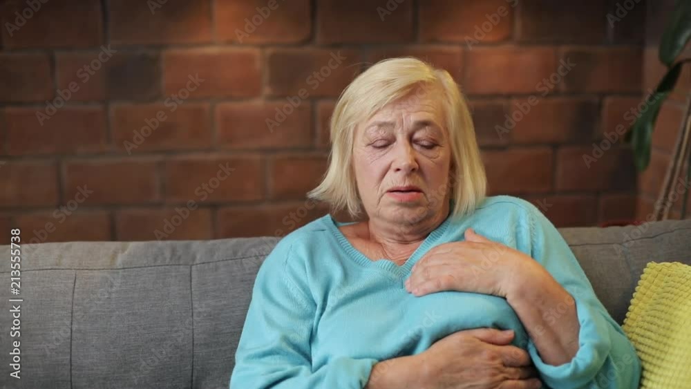 Nice grandmother laying on sofa, touching heart. Caucasian woman aged wearing blue sweater having heartache. Indoors. Disease. Living room.