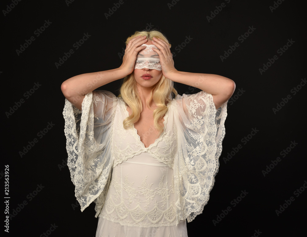 portrait of blonde girl wearing a white lace dress and blindfold.  black studio background.
