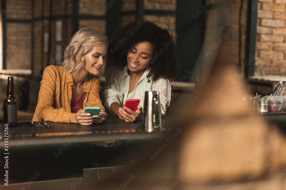 Fototapeta premium Interesting content. Beautiful young women sitting at the bar counter and looking through social media timelines while showing each other the most interesting posts
