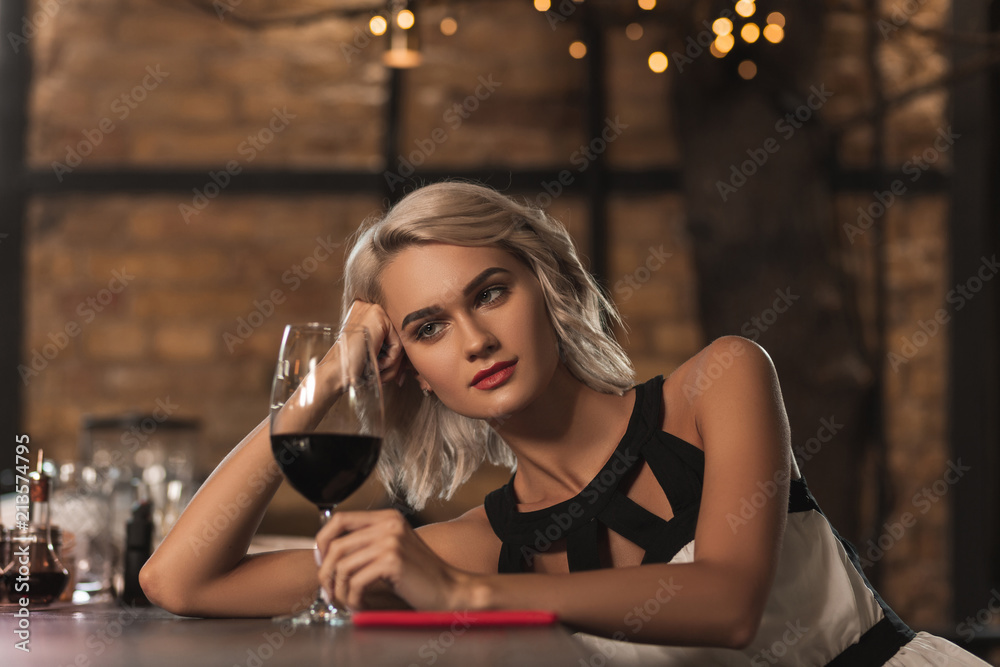 Good wine. Beautiful young blonde woman sitting at the bar counter and looking at her glass of wine while thinking about something