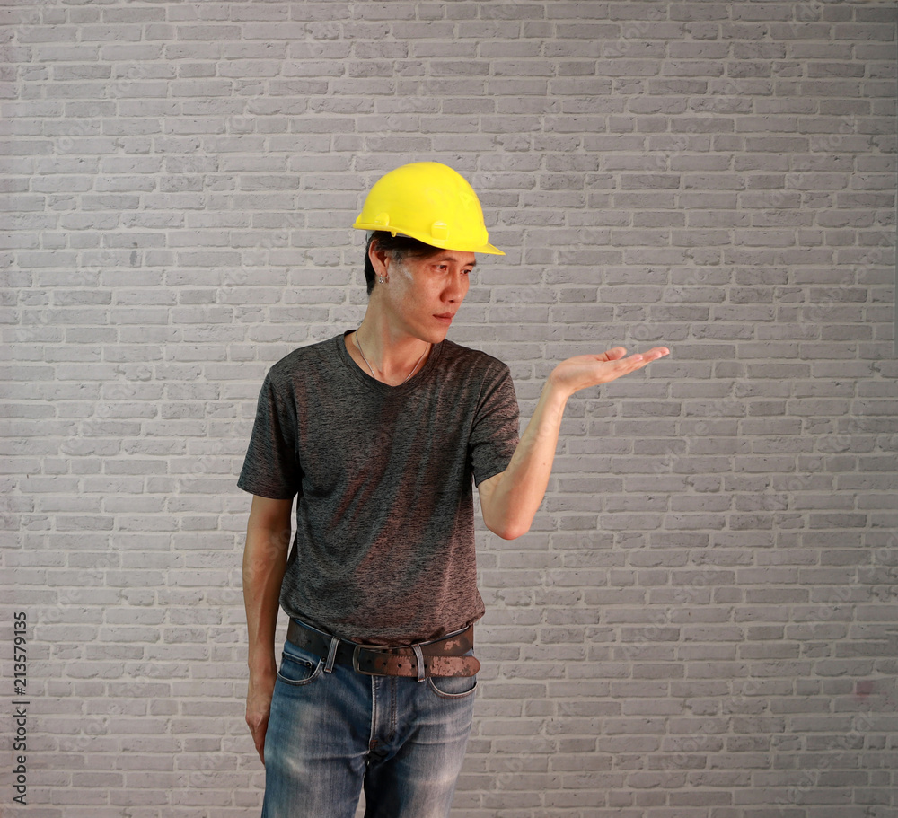 technician man ware yellow helmet with dark grey T-shirt and denim jeans standing and palms facing up of left hand on grey brick pattern background. technician man ware yellow helmet with dark grey T-shirt and denim jeans standing and palms facing up of left hand on grey brick pattern background.
