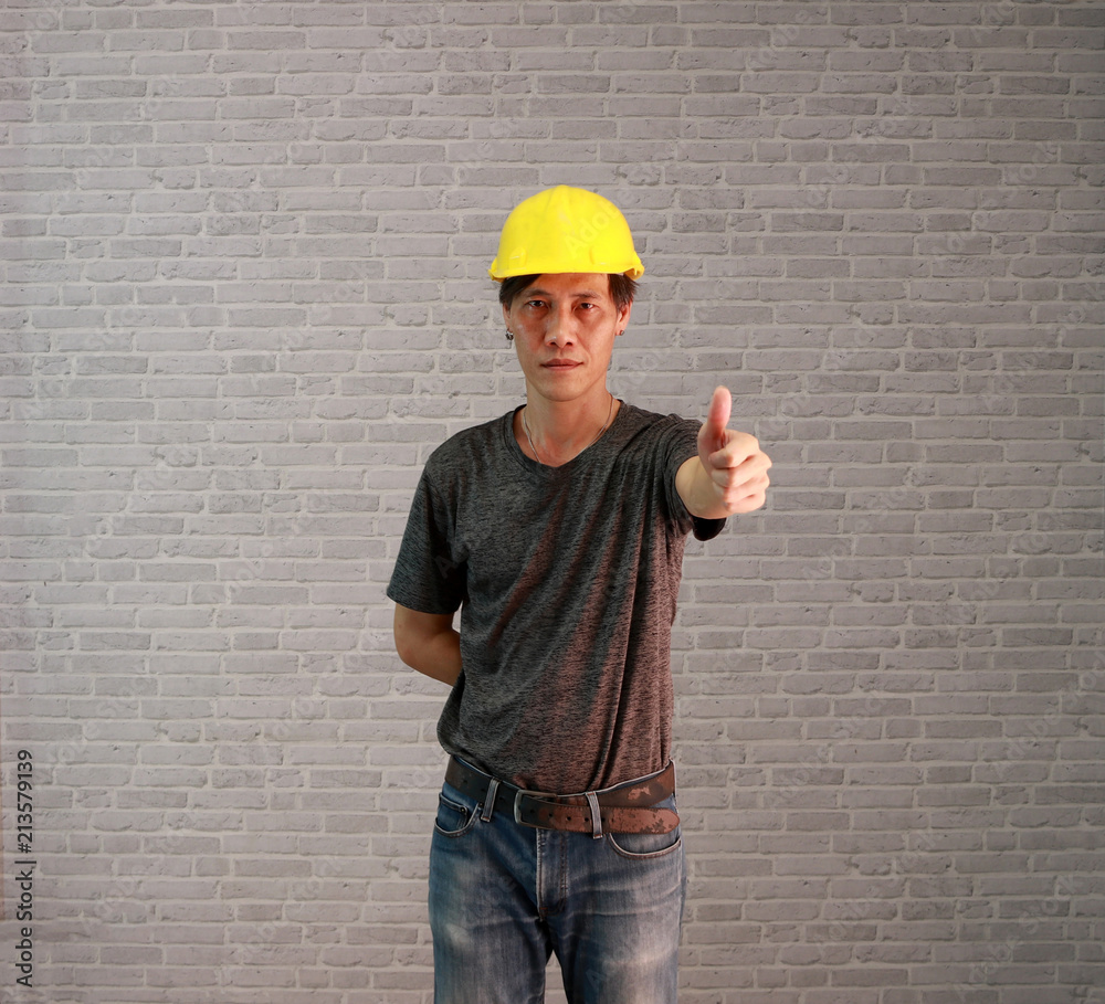 Technician man ware yellow helmet with dark grey T-shirt and denim jeans standing and thumbs up on grey brick pattern background. Technician man ware yellow helmet with dark grey T-shirt and denim jeans standing and thumbs up on grey brick pattern background.