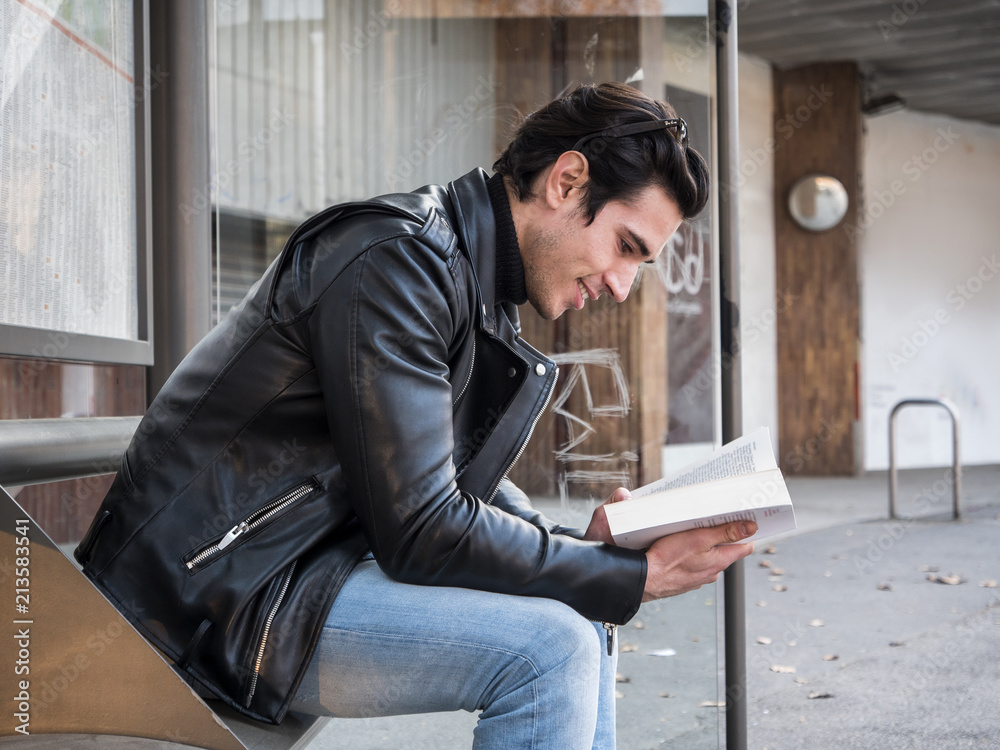 Side view of handsome stylish man in leather jacket sitting on bench on  street and reading book with smile Stock Photo | Adobe Stock