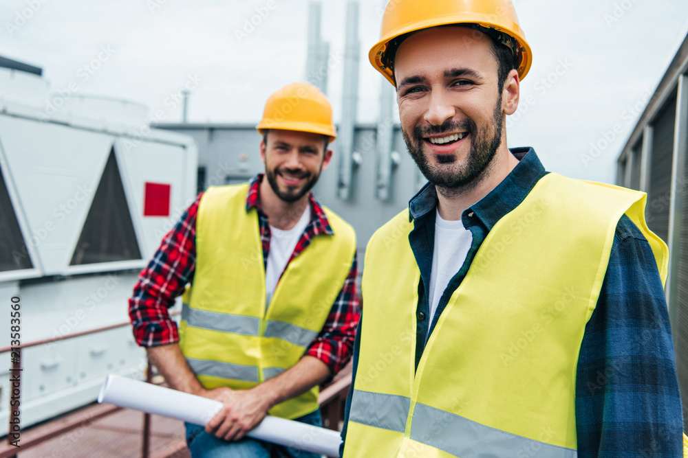 smiling engineers in safety vests and helmets with blueprint on roof