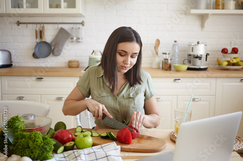 Portrait of attractive casually dressed young dark haired chubby woman cutting vegetables using sharp and cooking board  sitting at kitchen table  watching video recipe online on laptop computer