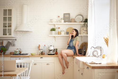 Portrait of cute brunette girl eating delicious pastry, sitting alone on counter in modern kitchen interior. Playful funny barefooted young female with dark hair having doughnut for breakfast