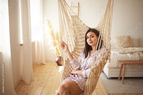 Indoor portrait of beautiful young brunette plus size woman wearing silk night shirt with stripes swinging in hanging woven chair in cozy bedroom interior, looking and smiling happily at camera photo