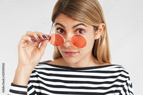 People, style, fashion and accessories concept. Cute bug eyed young blonde female model wearing striped top staring at camera surprisingly, holding pink round sunglasses, advertising optics