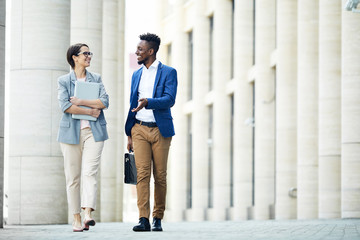 Happy excited office colleagues in stylish outfits talking about work when walking over street in summer