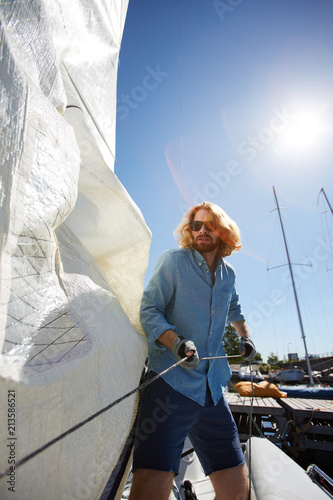 Serious handsome hipster guy with long hair and beard wearing sunglasses and gloves pulling rope while adjusting sail before sailing in summer