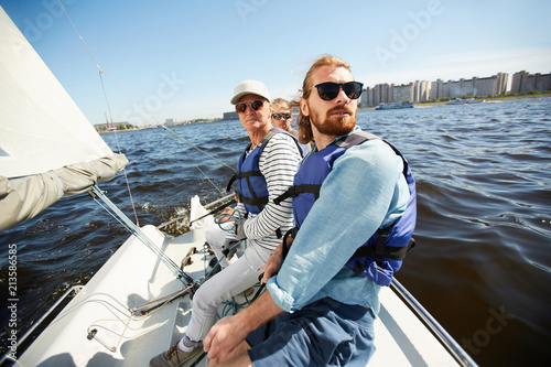Serious calm men in life jackets and sunglasses sitting in row on yacht and looking around during sailing tour on river