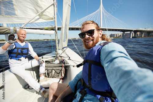 Cheerful handsome friends in blue life jackets taking selfie against beautiful bridge while sitting on boat deck
