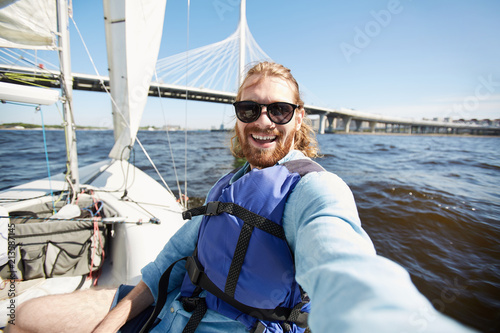 Happy excited handsome young man with beard wearing sunglasses laughing while photographing himself against urban bridge while floating on yacht