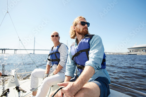 Pensive dreamy male tourists in sunglasses wearing life jackets sitting on sailboat deck and looking around during sailing tour © pressmaster
